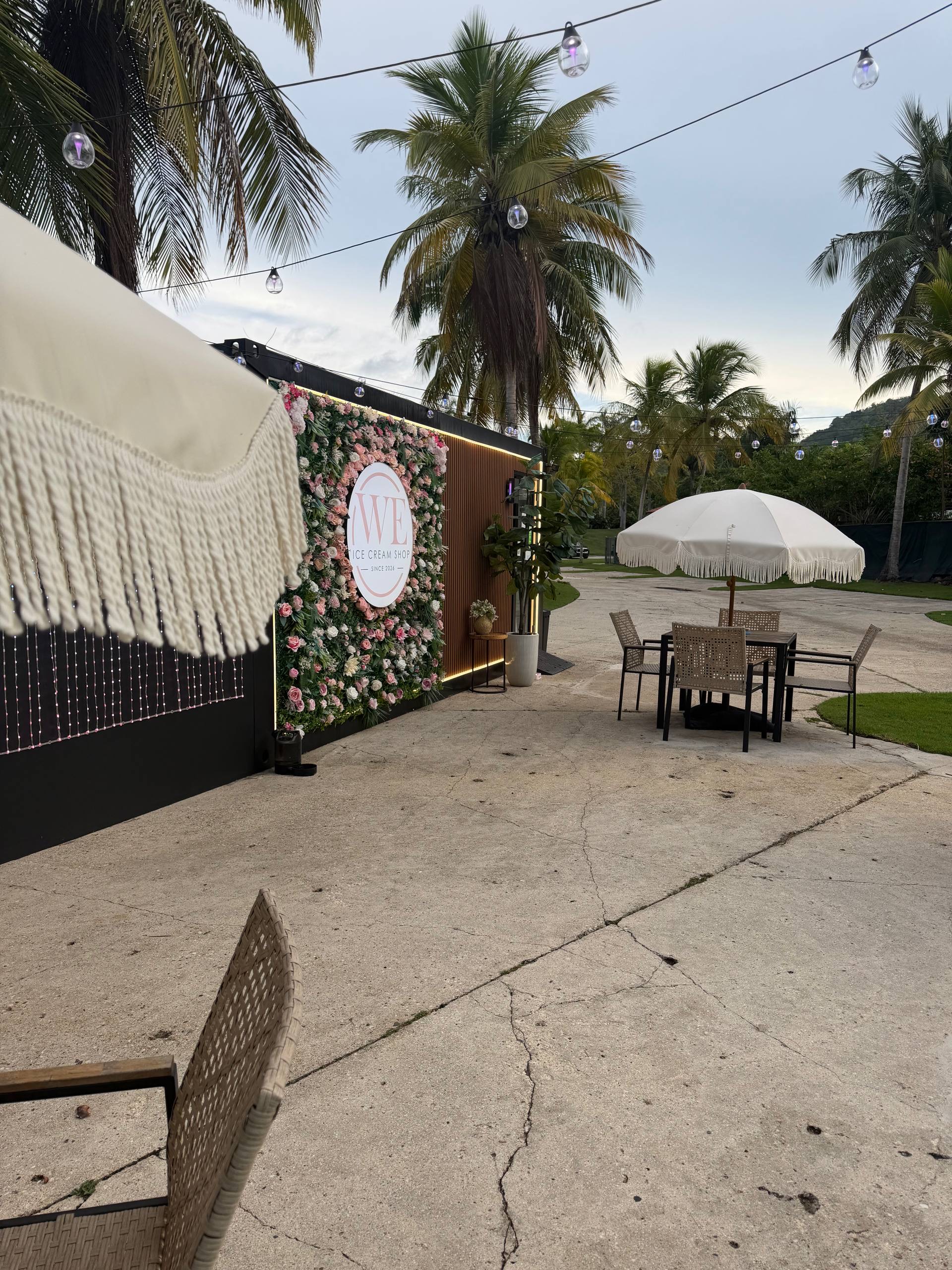 Patio de WE Ice Cream Shop por la tarde, con el mural de flores, sombrillas y luces colgantes